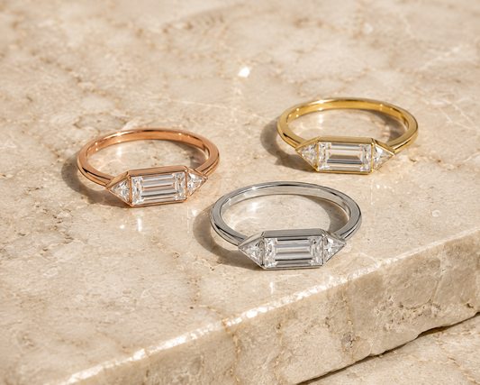 Emerald-cut three-stone moissanite rings in silver, yellow, and rose-gold tones on beige stone